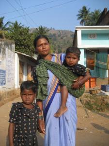 Mother carries younger child in sling while older child walks with her.